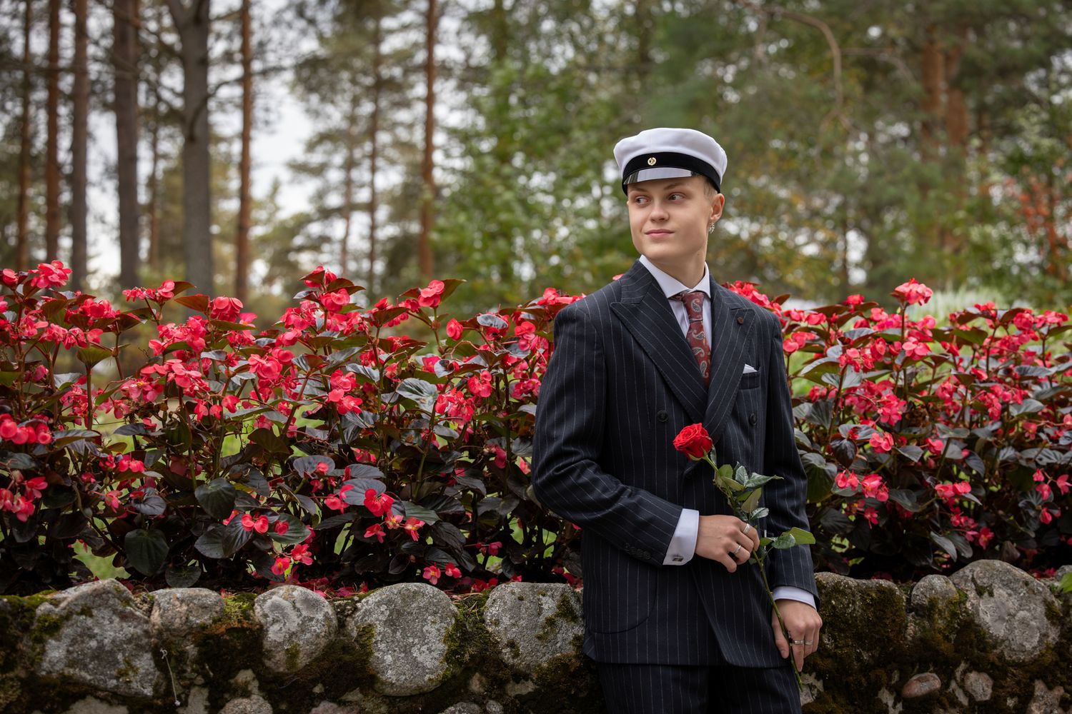 Graduate in cap and gown stands in front of vibrant red azalea bushes during spring commencement ceremony.