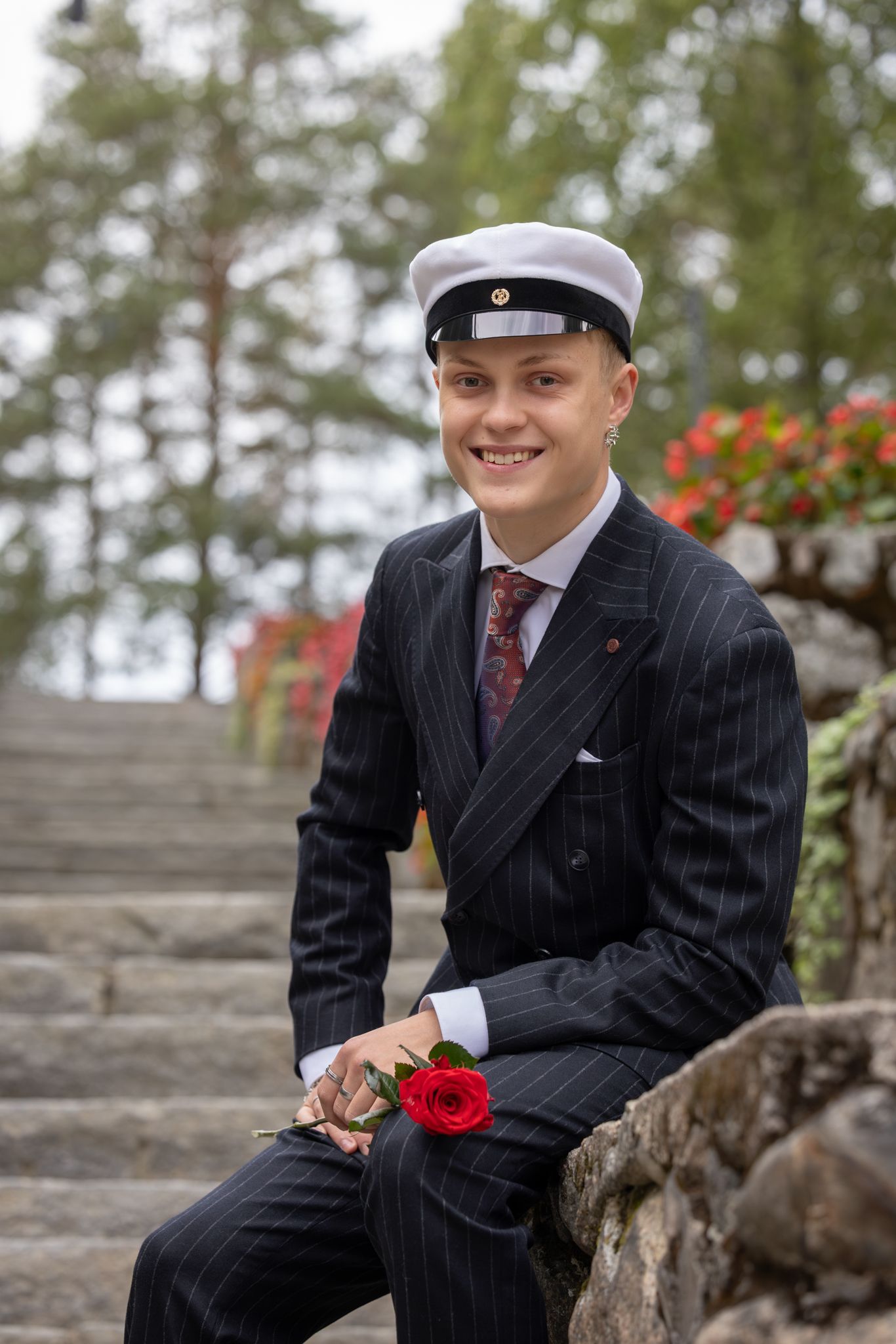 A person in a pinstripe suit and sailor cap sits on stone steps holding a red rose.