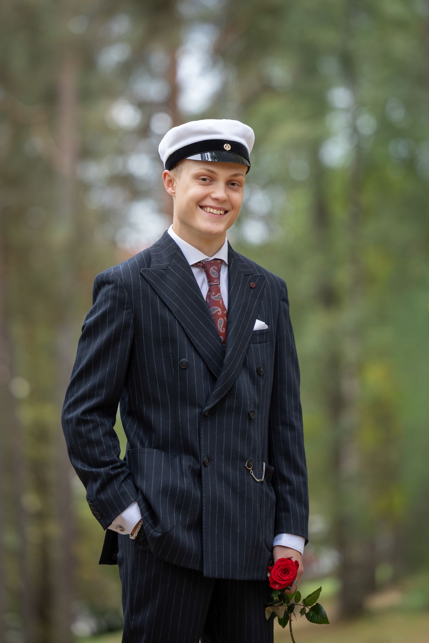 A person in a pinstripe suit and cap poses outdoors with a red rose against a blurred forest background.