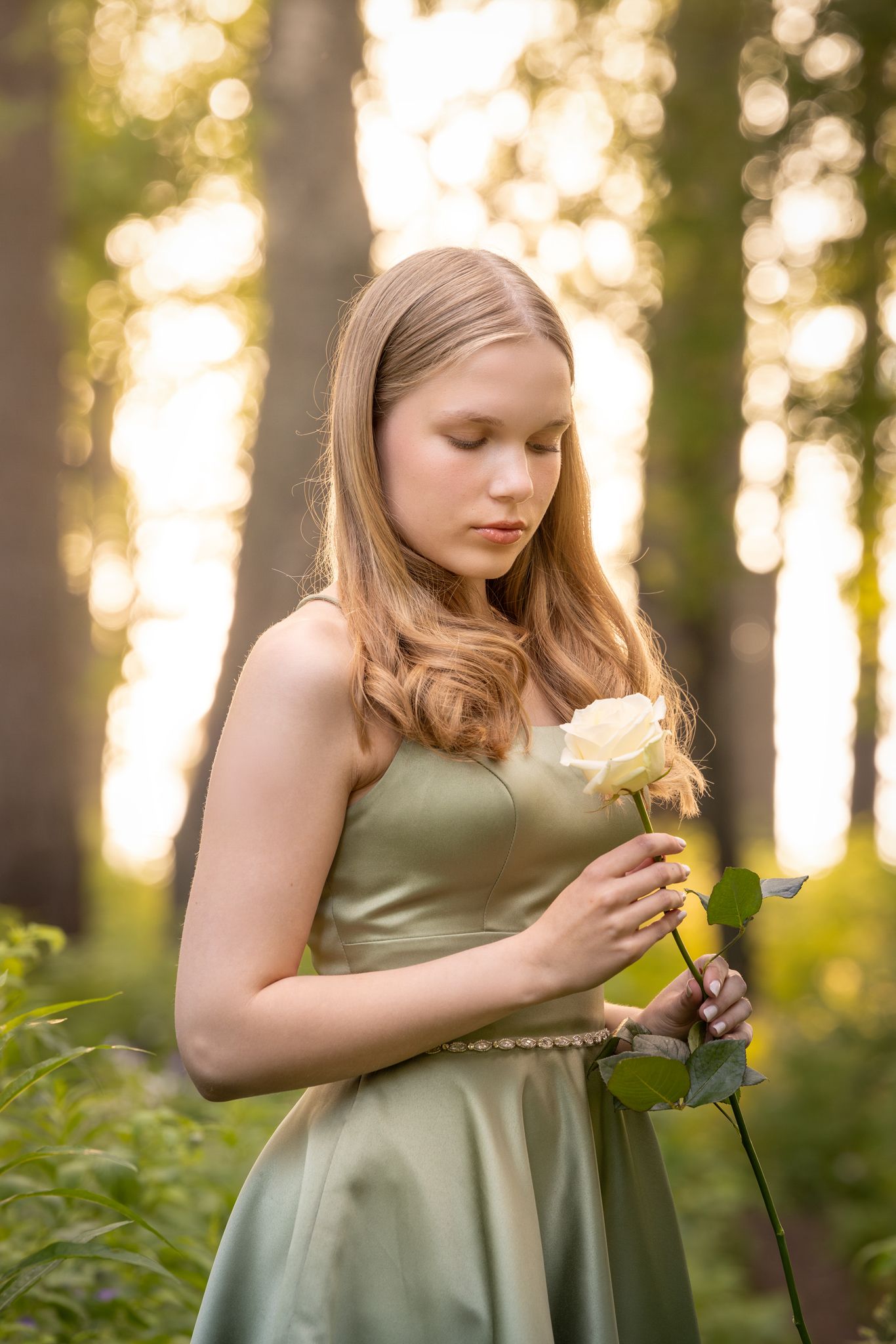 A dreamy golden-hour scene in a forest with someone in a sage green dress delicately examining flowers.
