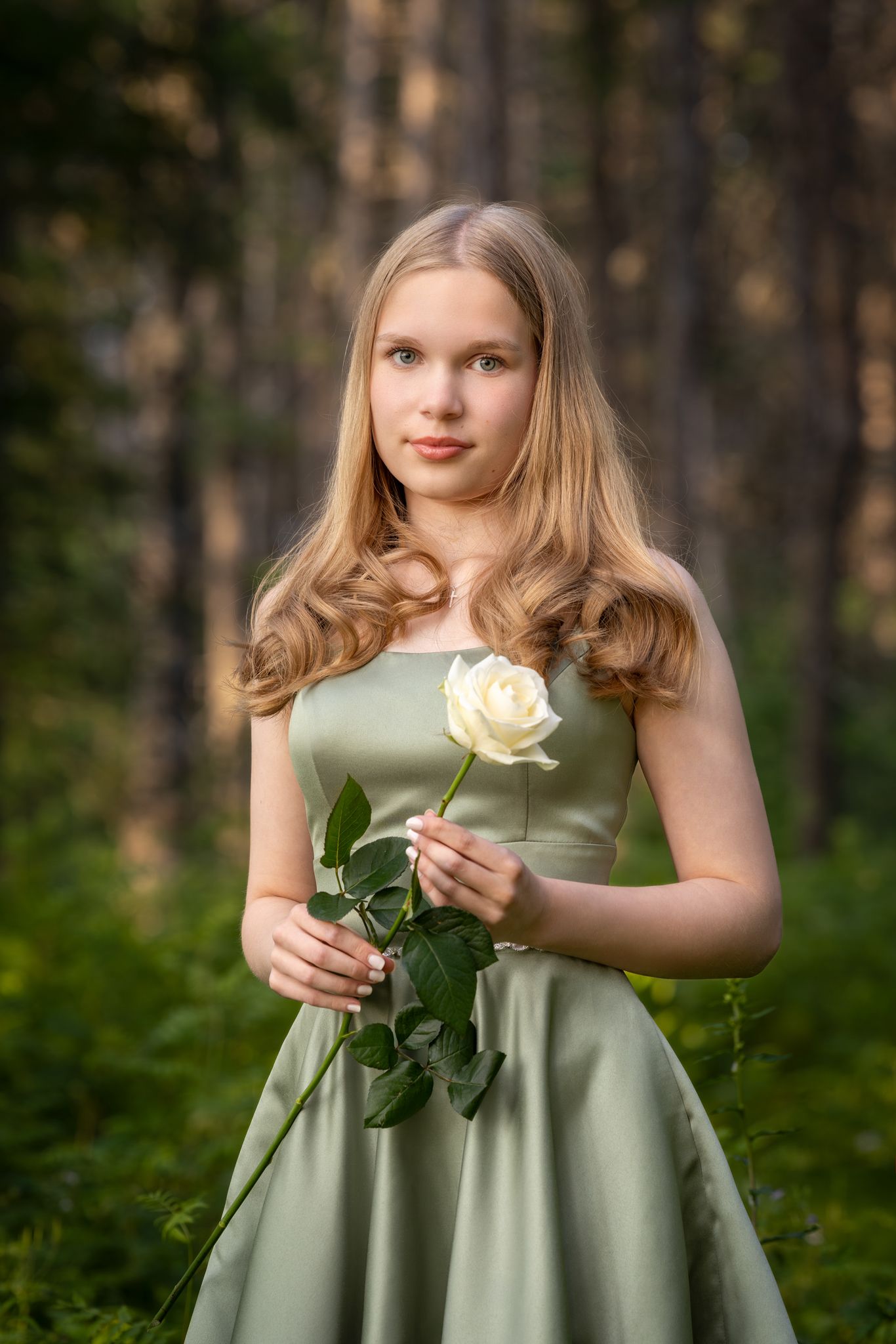 Young woman in sage green dress holds white rose while standing in wooded setting during golden hour.