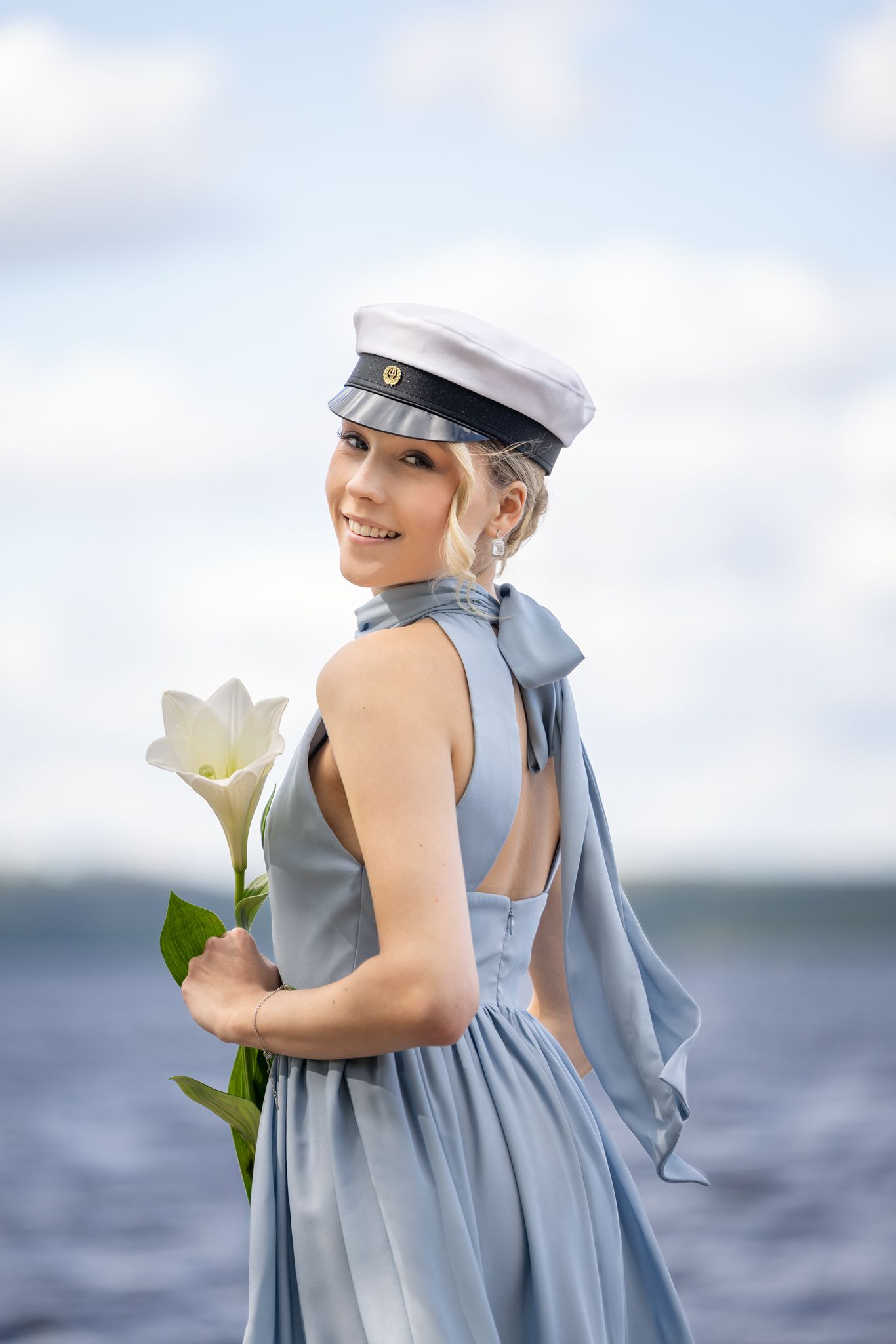 Young woman in light blue dress and white captain hat holds a white lily flower by the sea.
