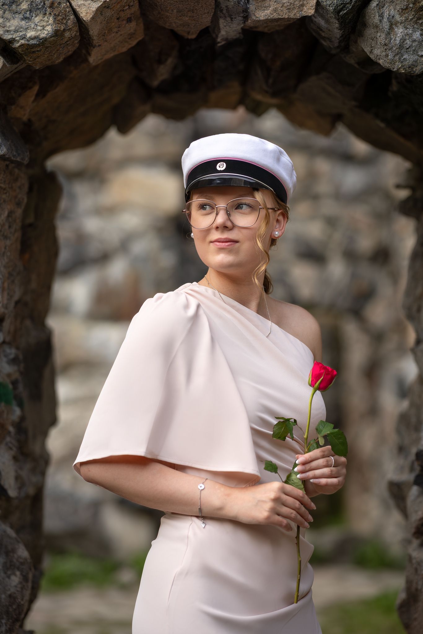 Young woman in white cap and cream dress holding a red rose, standing under a stone arch outdoors.
