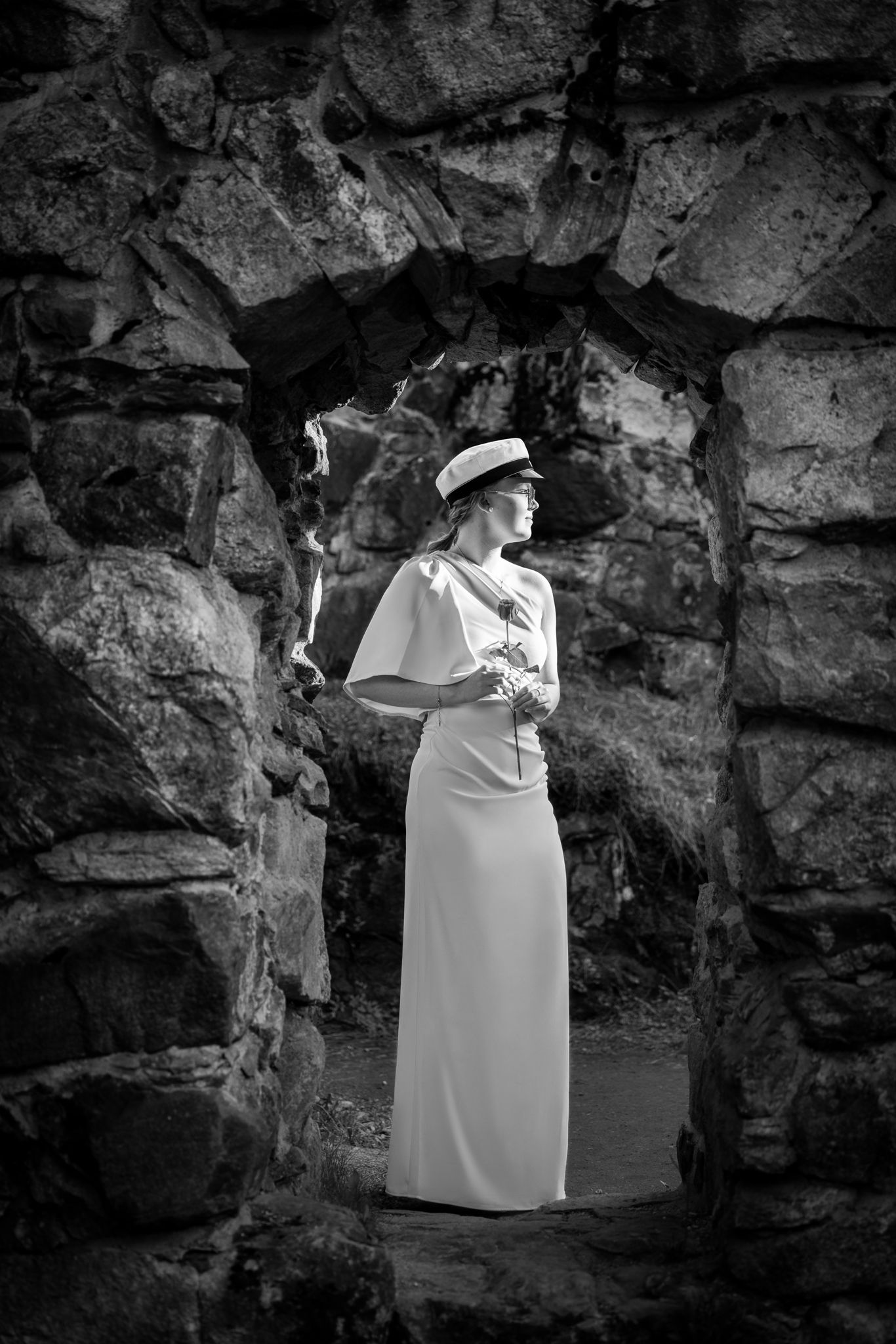 Black and white photo of elegant woman in white gown and hat standing in stone archway ruins.