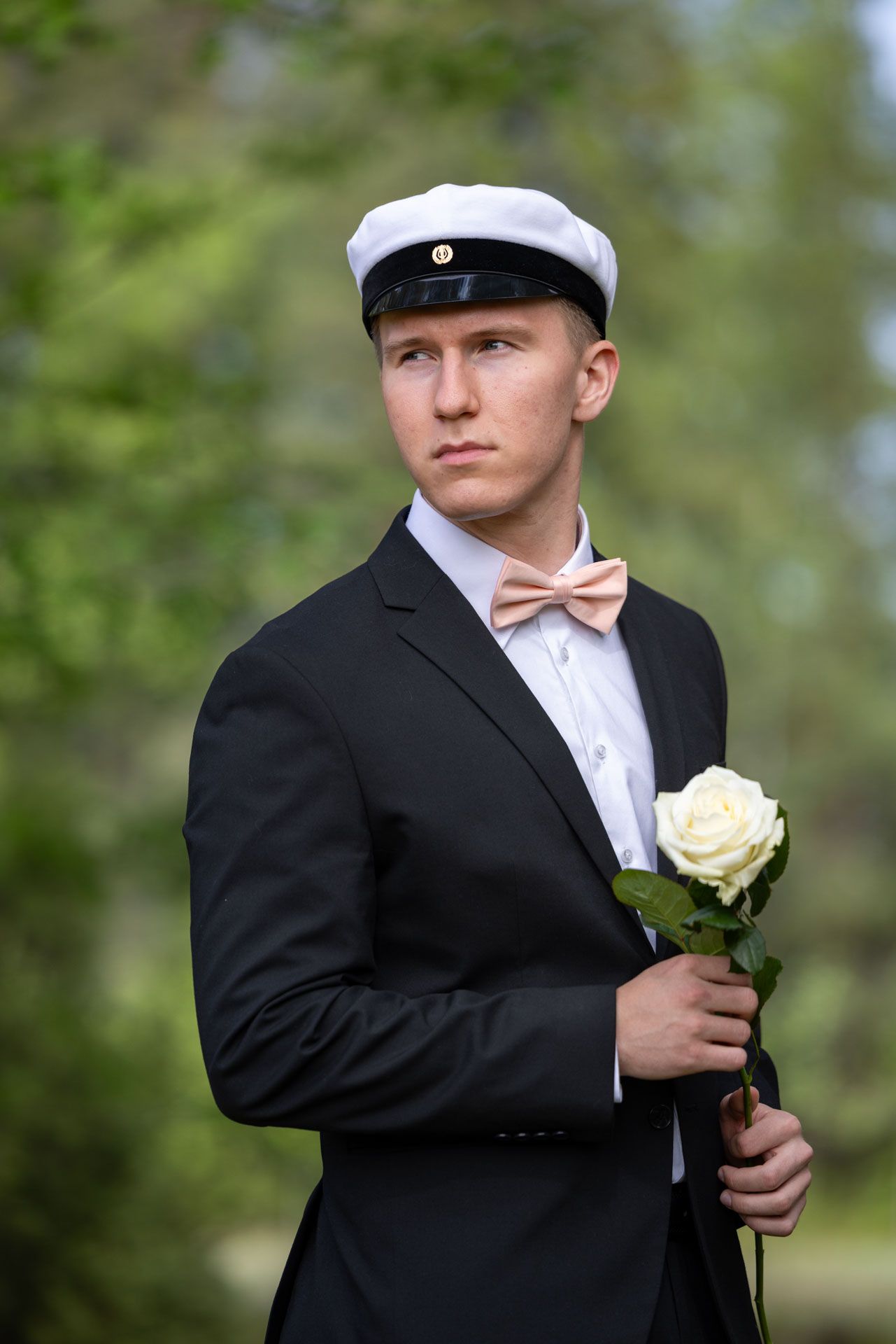 Young man in black suit, bow tie, and white cap holding a white rose outdoors.