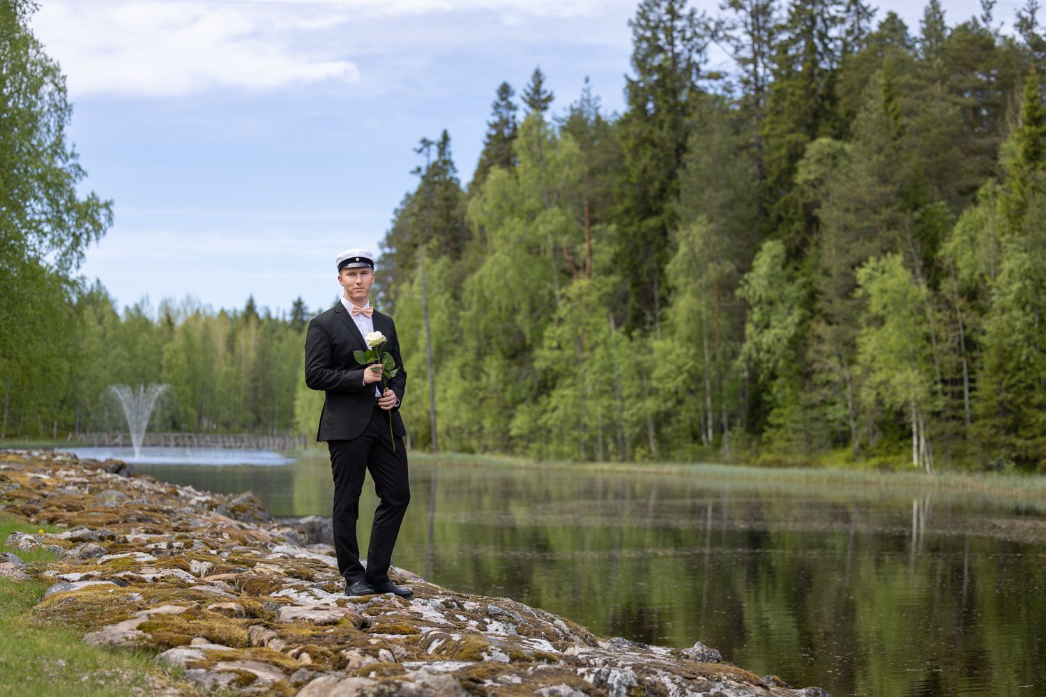 Man in black suit holding flowers stands on rocky shore beside a calm lake surrounded by lush green forest.