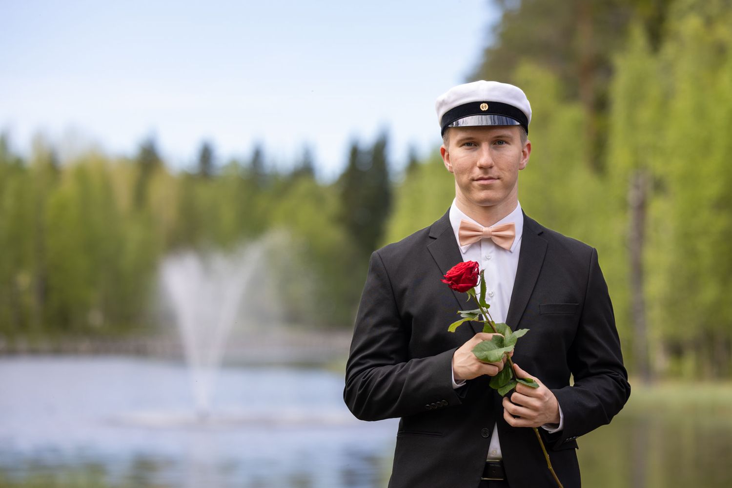 Young man in formal suit and white cap holding a red rose near a fountain outdoors.