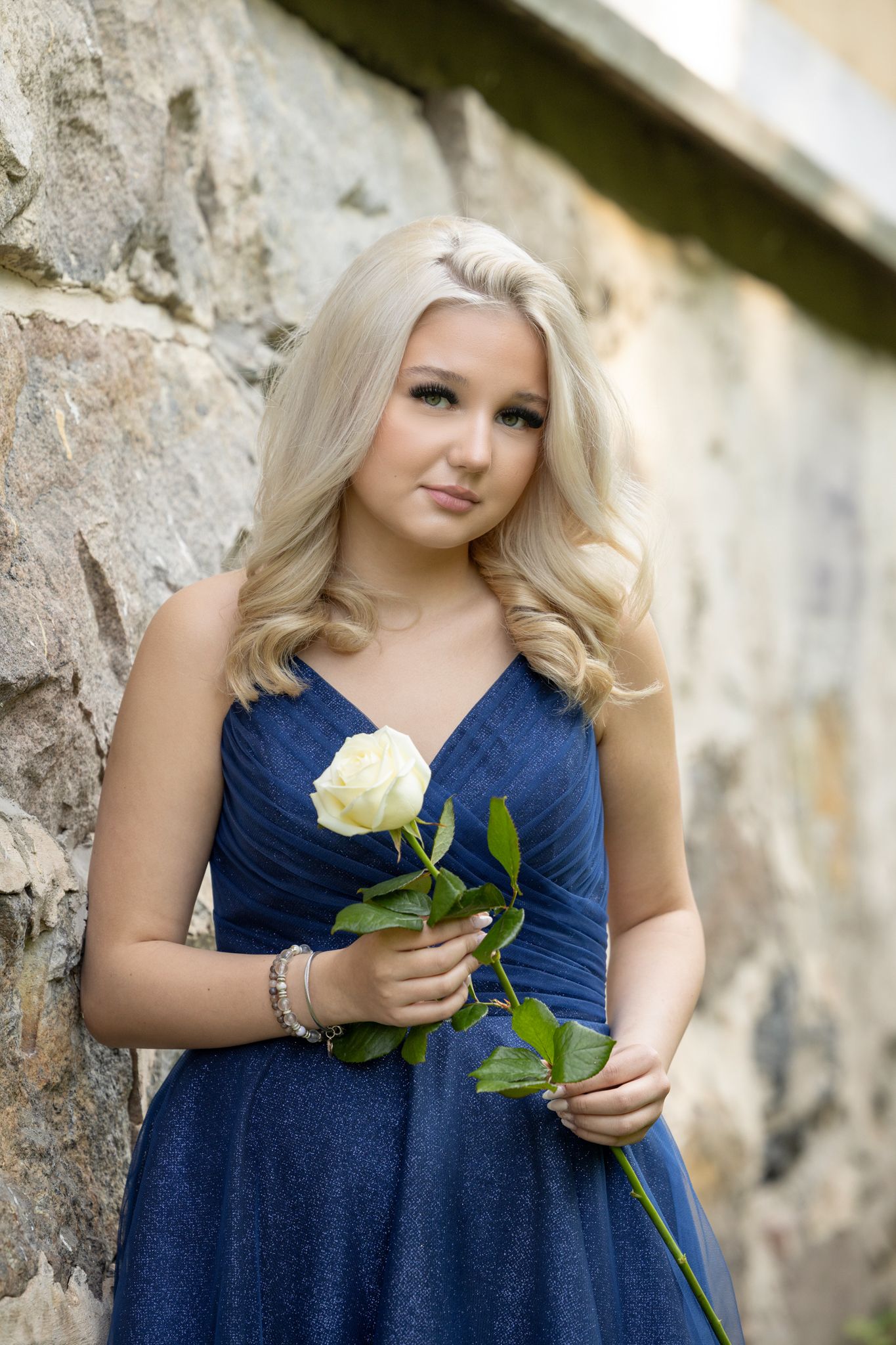 Young woman in navy blue dress holds a single white rose against stone wall.