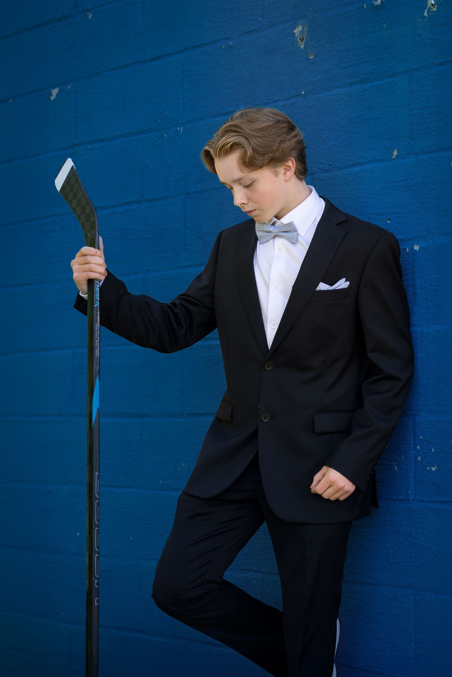 Young man in black tuxedo with bow tie holds hockey stick against blue wall.