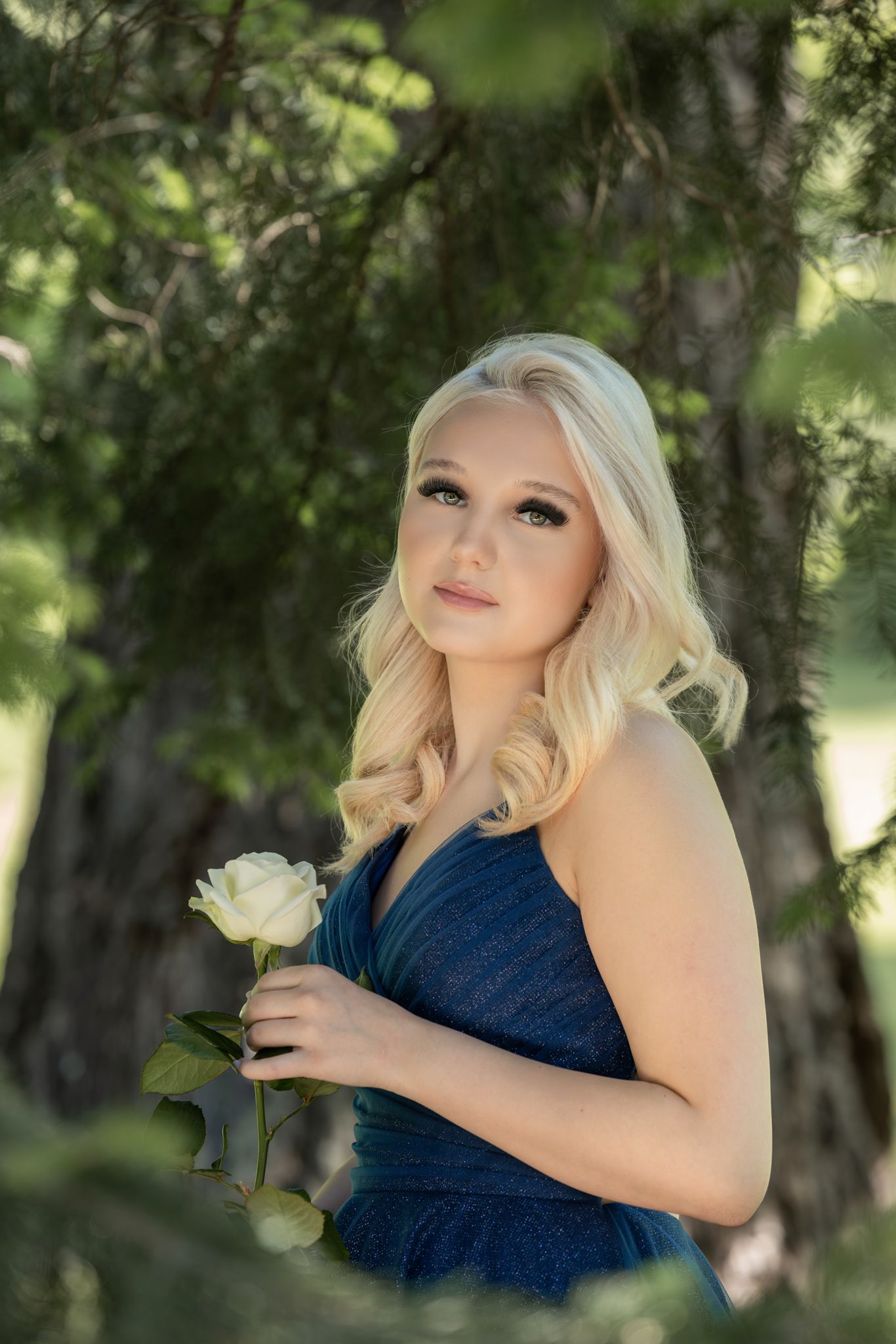 A young woman in a navy blue dress poses outdoors with a white rose against a blurred natural background.