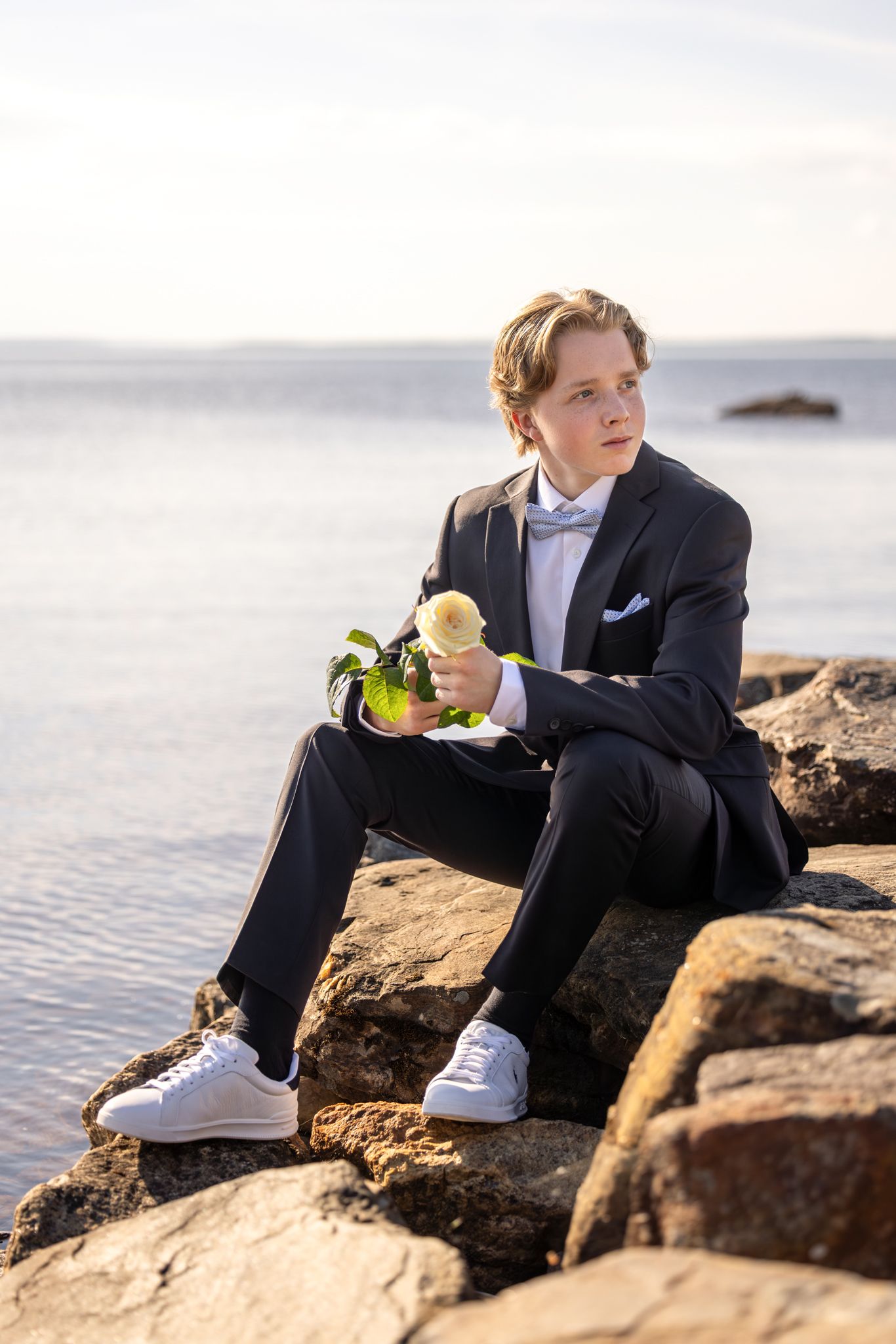 Young boy in formal black suit holding a white rose, sitting on rocky shoreline with calm water in background.