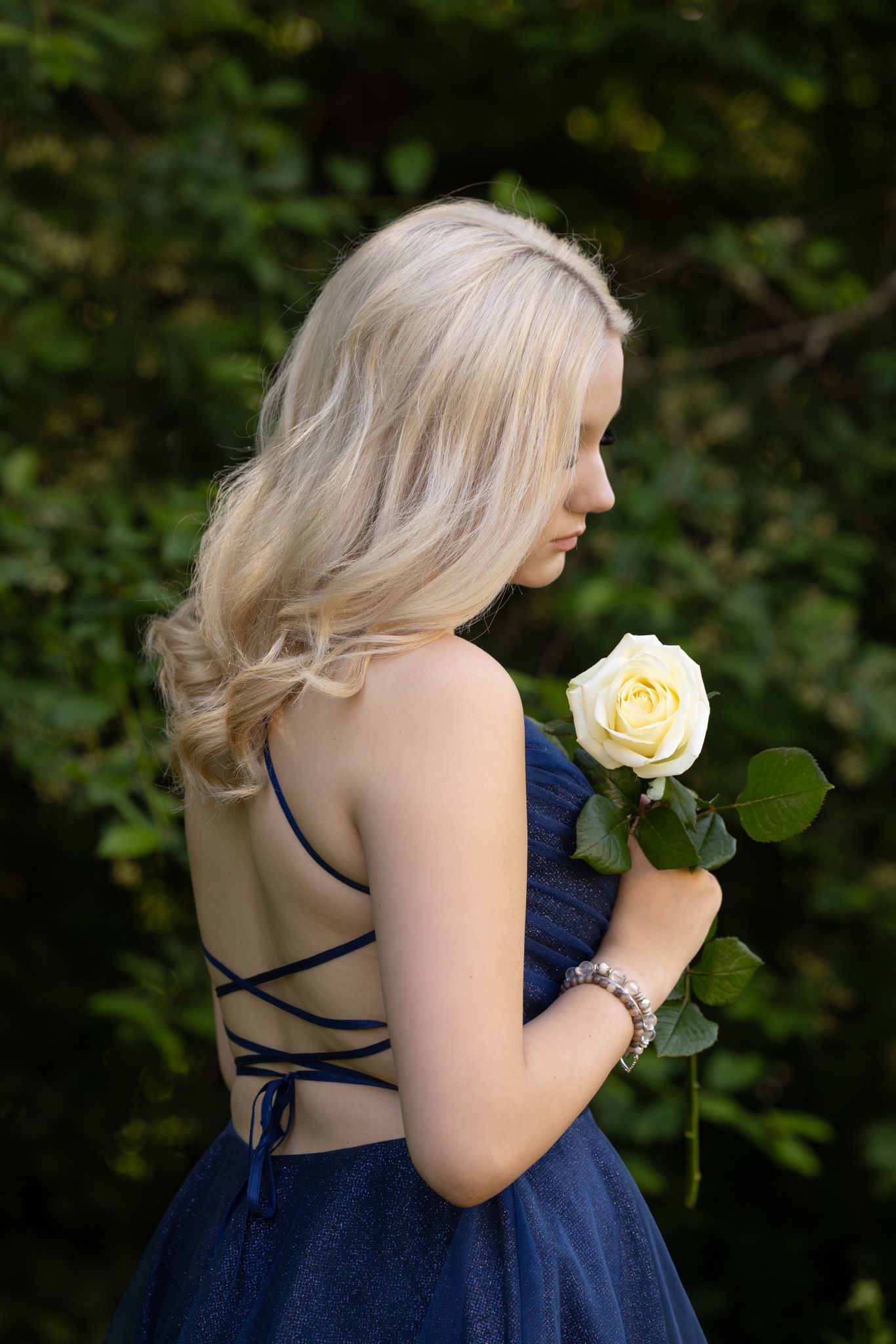 A person in a navy blue backless dress holds a single cream-colored rose against a garden backdrop.