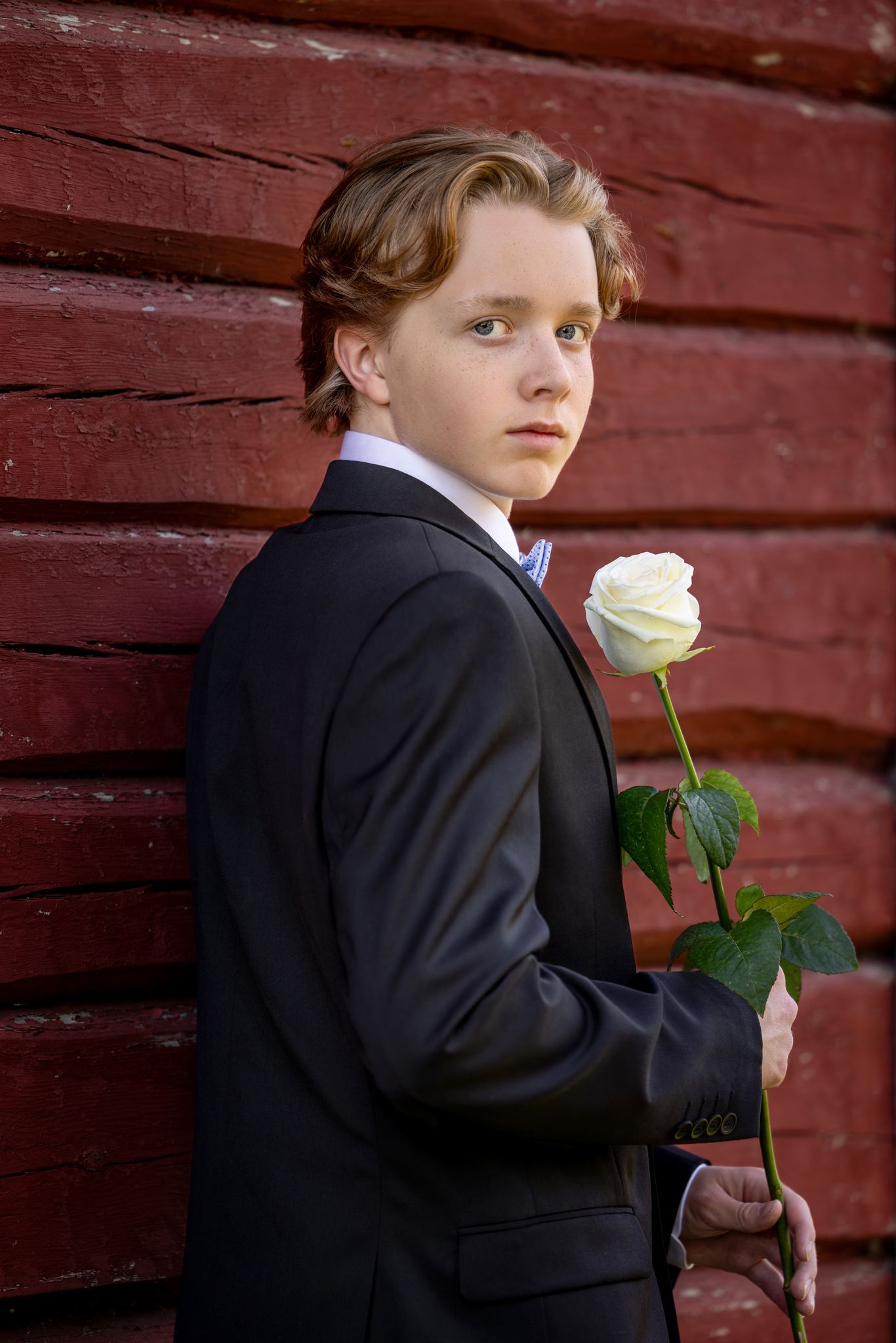 Young man in black suit holding a white rose against a rustic red wooden wall backdrop.