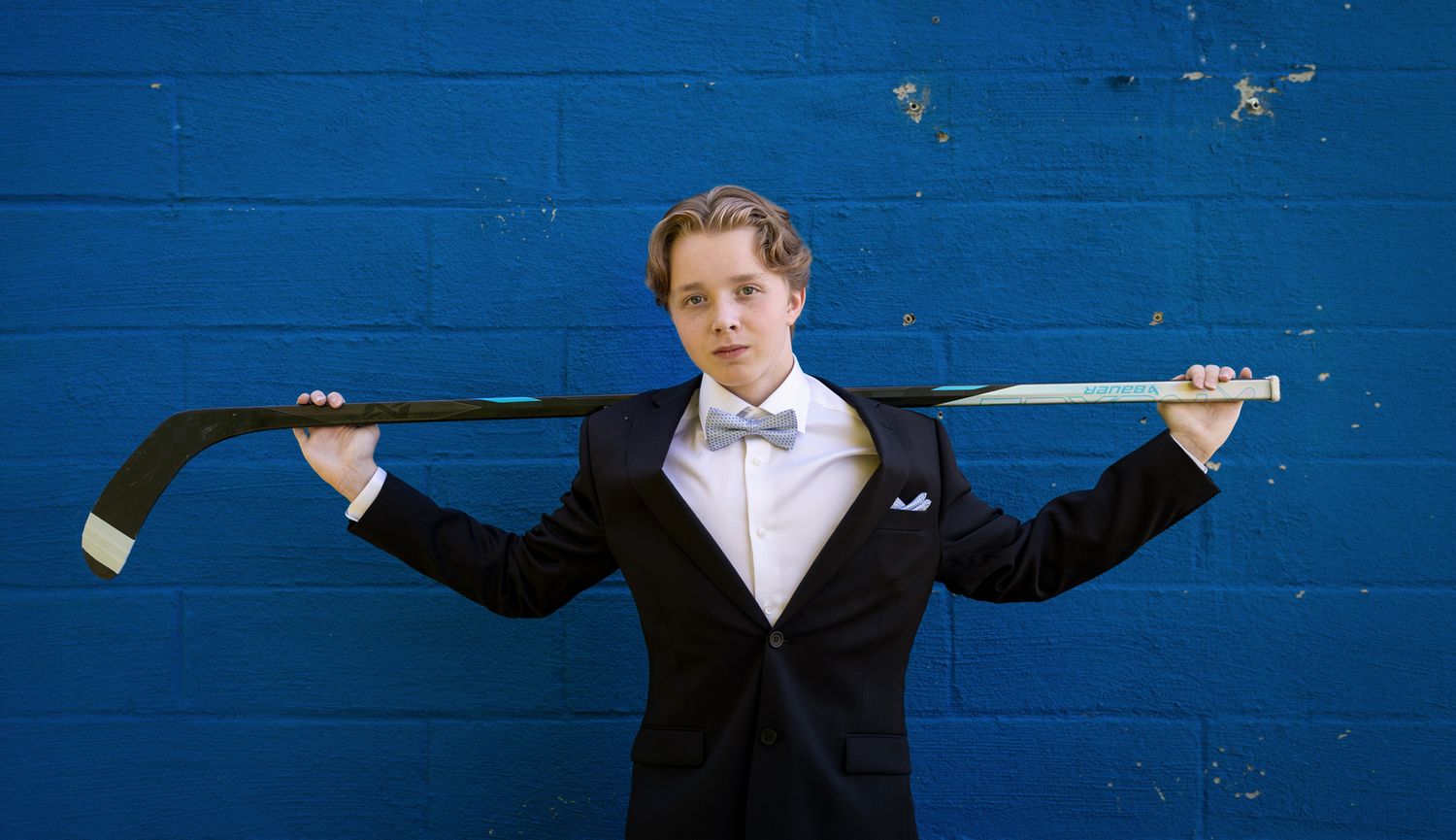 Young man in tuxedo and bow tie holding hockey stick across shoulders against blue wall.