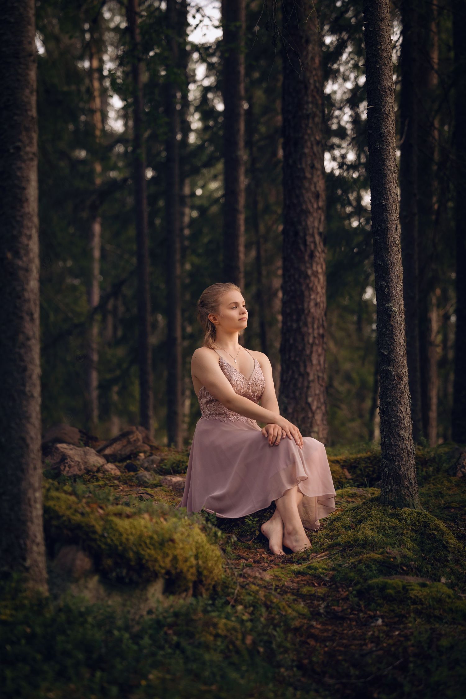 Vaaleanpunaiseen pukeutunut nuori nainen istuu sammaleisen metsän keskellä kannolla.