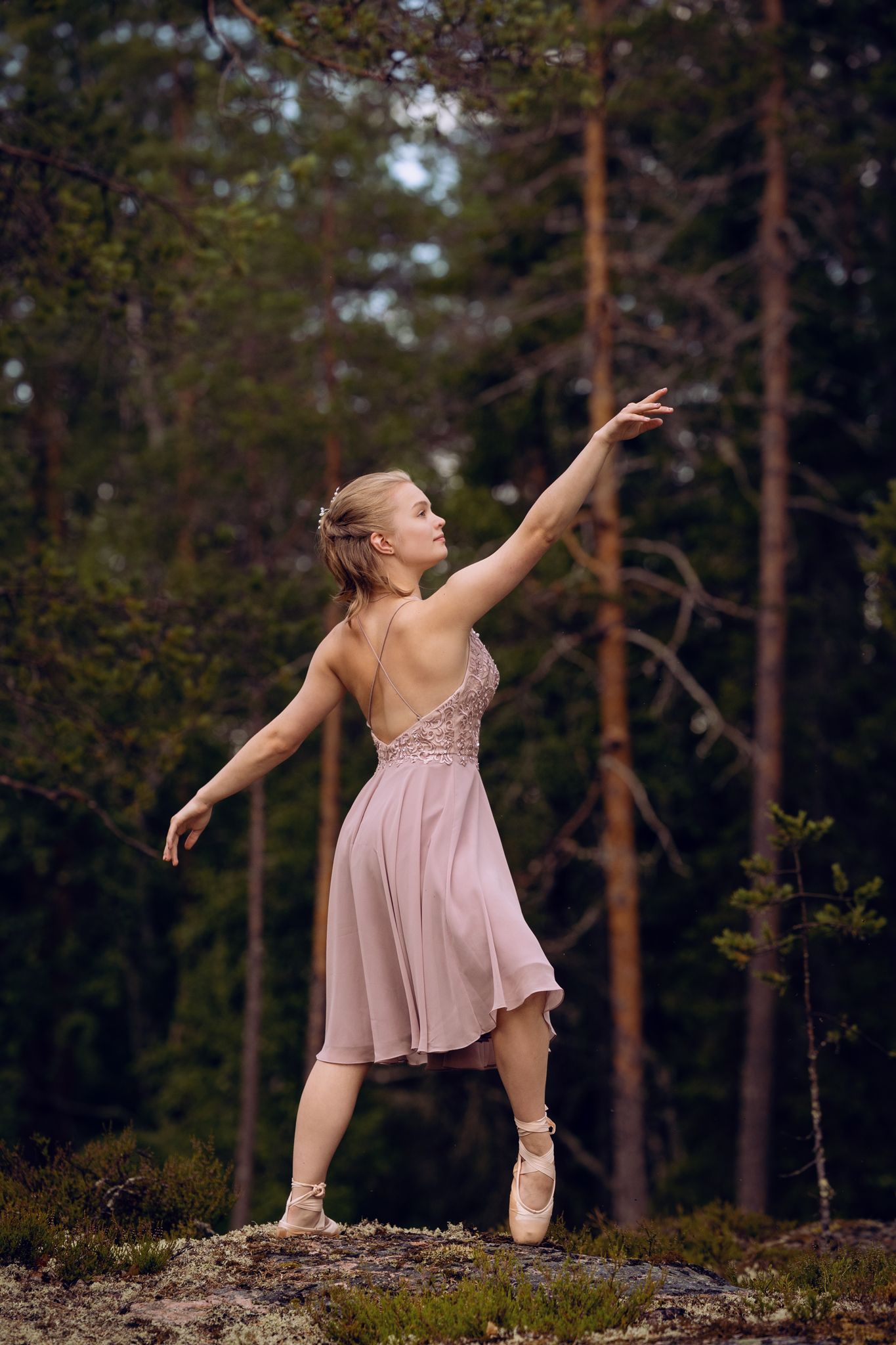 Vaaleanpunaiseen pukeutunut nuori nainen tanssii balettia metsässä. Taustalla puita.
