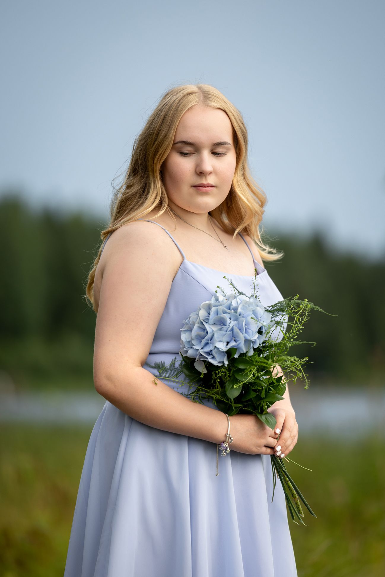 Siniseen mekkoon pukeutunut tyttö seisoo rantamaisemassa pitäen käsissään sinistä kukkaan.
