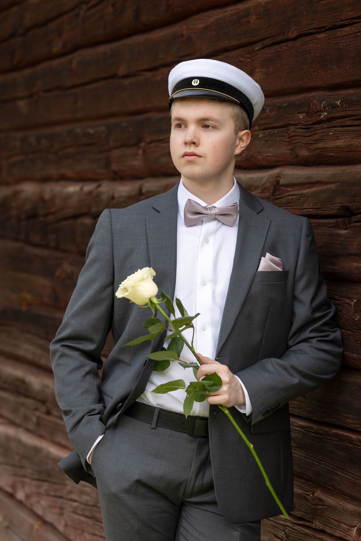 Harmaaseen pukuun pukeutunut ylioppilas nojaa punertavaan hirsiseinään, kädessään valkoinen ruusu.