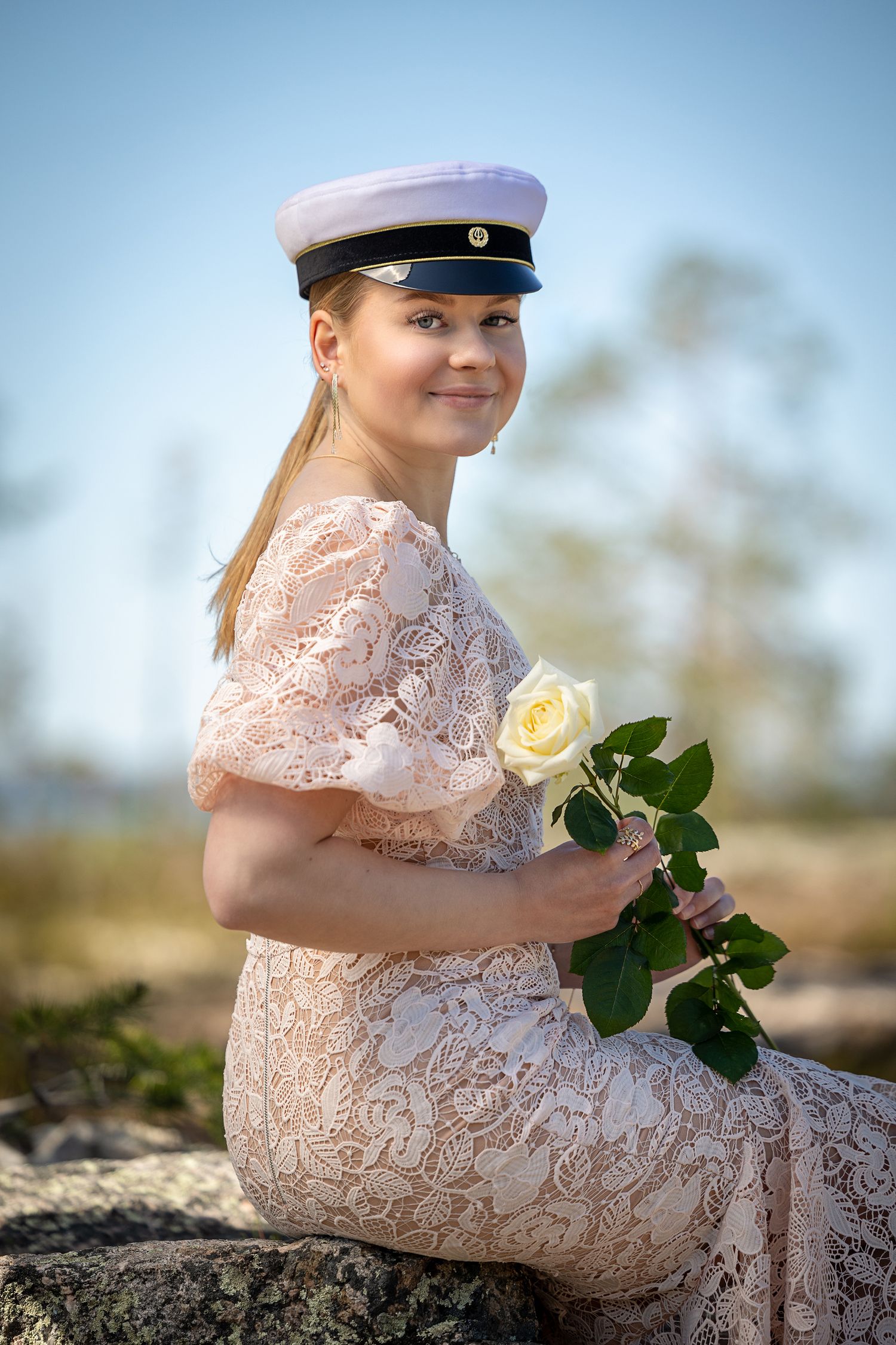 Vaaleanpunaiseen mekkoon pukeutunut ylioppilas istuu kivellä, käsisään valkoinen ruusu.