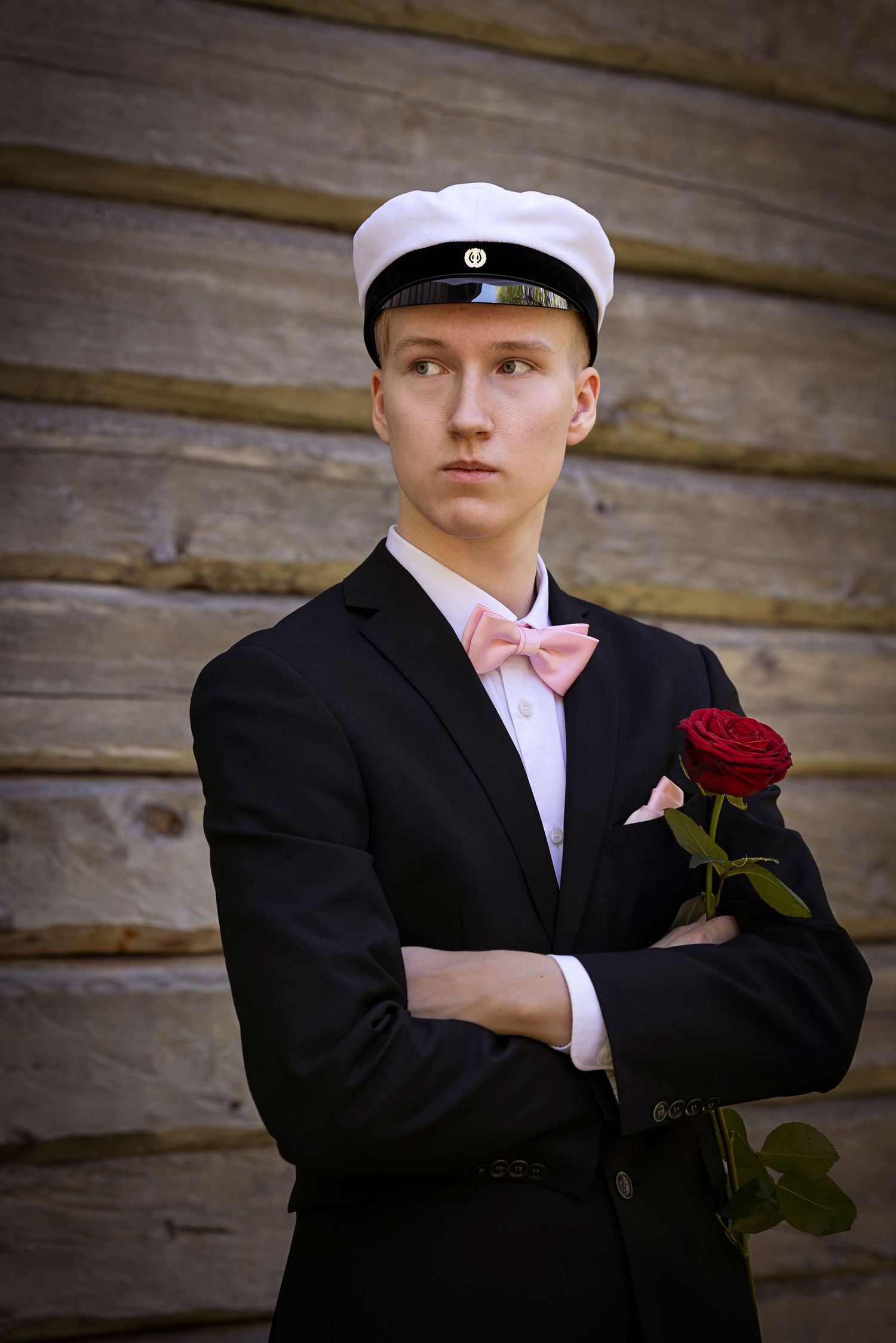 Mustaan pukuun ja vaaleanpunaiseen rusettiin pukeutunut ylioppilas kädet puuskassa ja käsissä punainen ruusu.