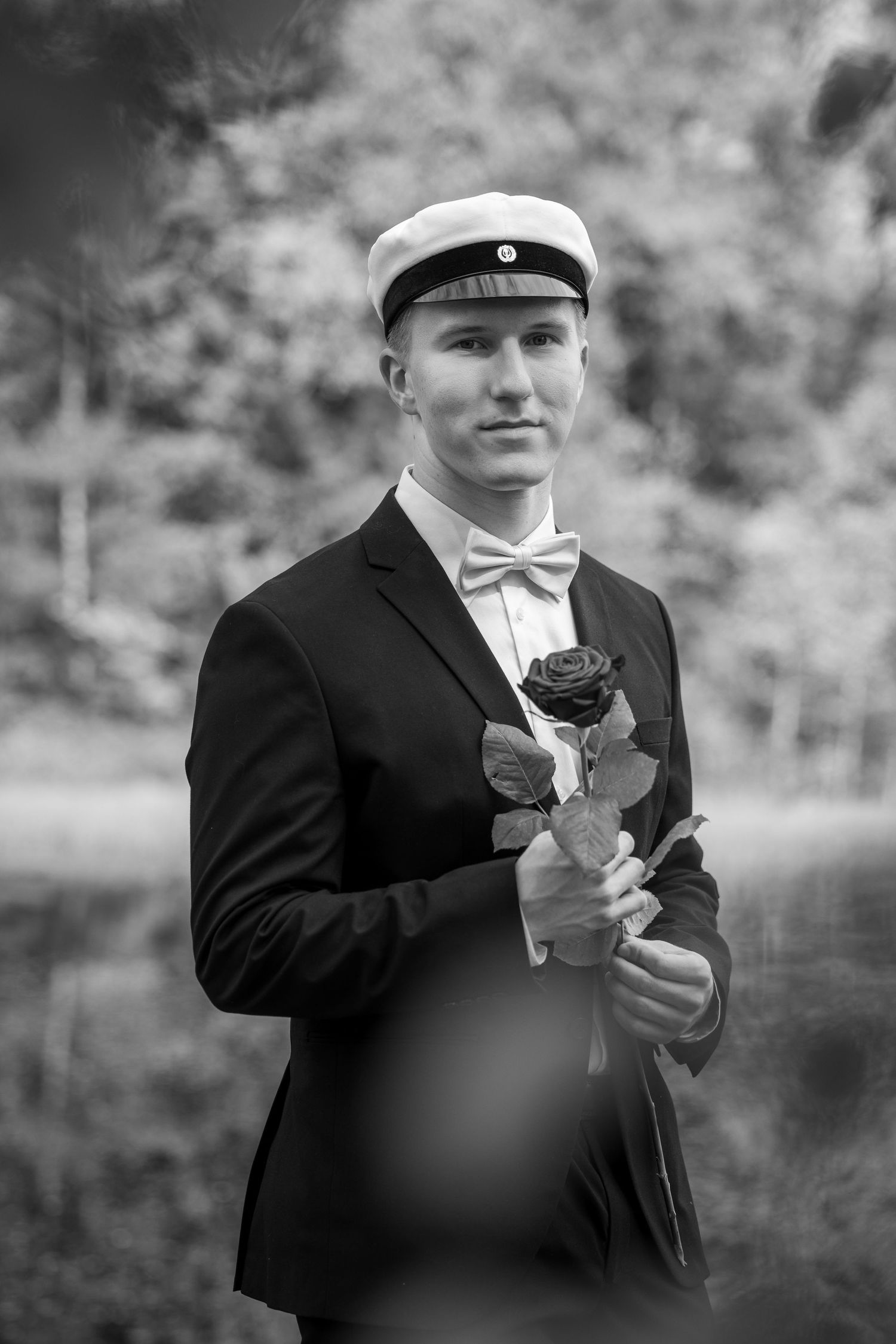 Young man in black suit, bow tie, and white cap holds a rose outdoors in a classic black and white portrait photo.