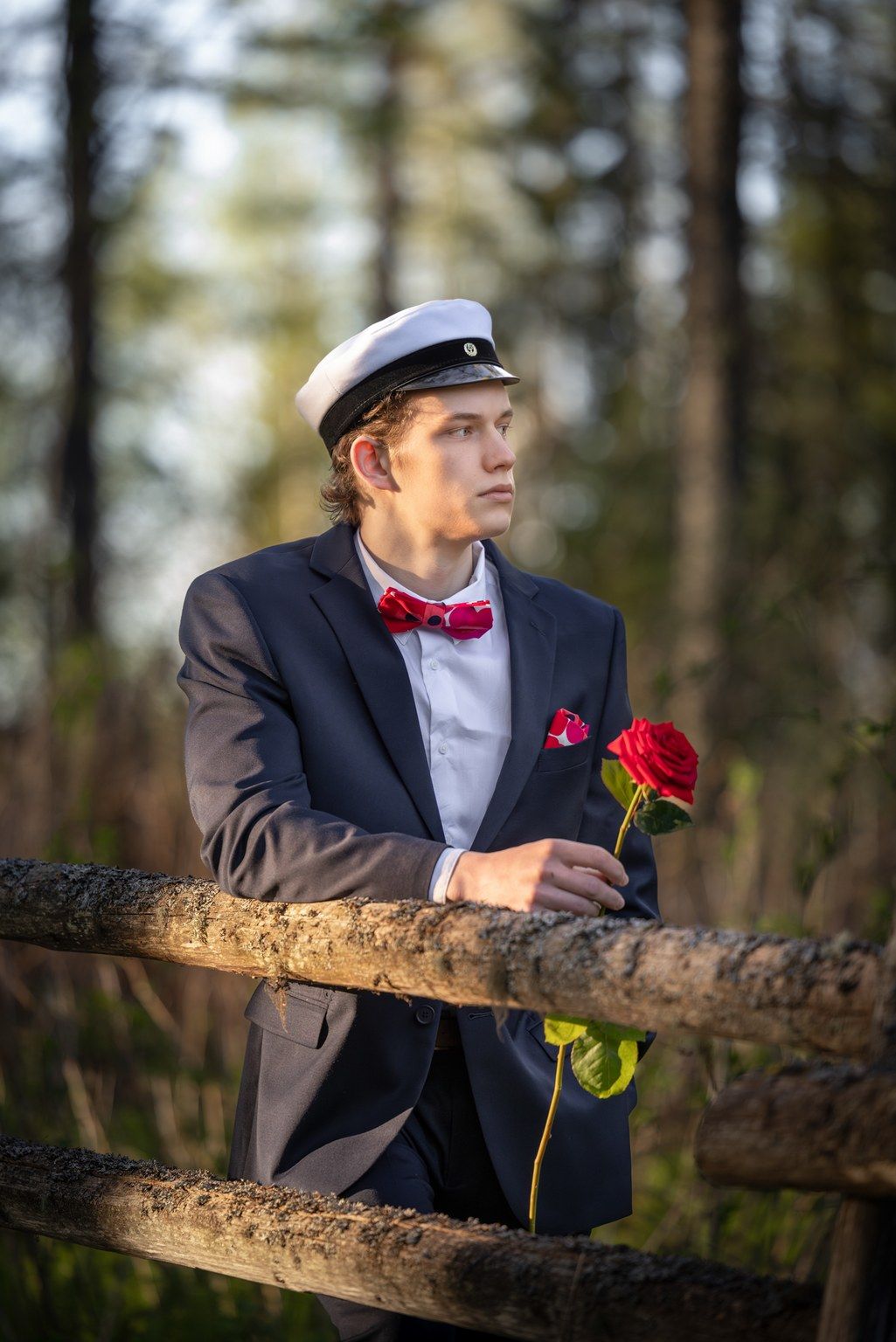 Tummansiniseen pukuun pukeutunut nuori ylioppilasmies nojaa aitaan käsissään punainen ruusu.