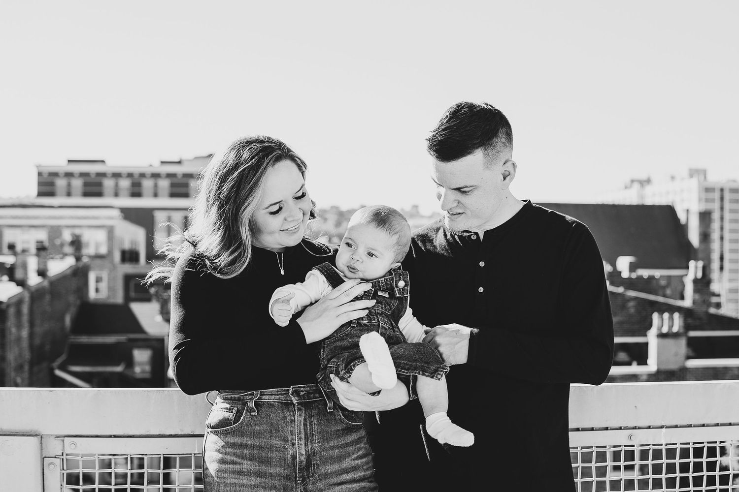 A couple stands on an urban rooftop with a baby, sharing intimate family moments in black and white photography.
