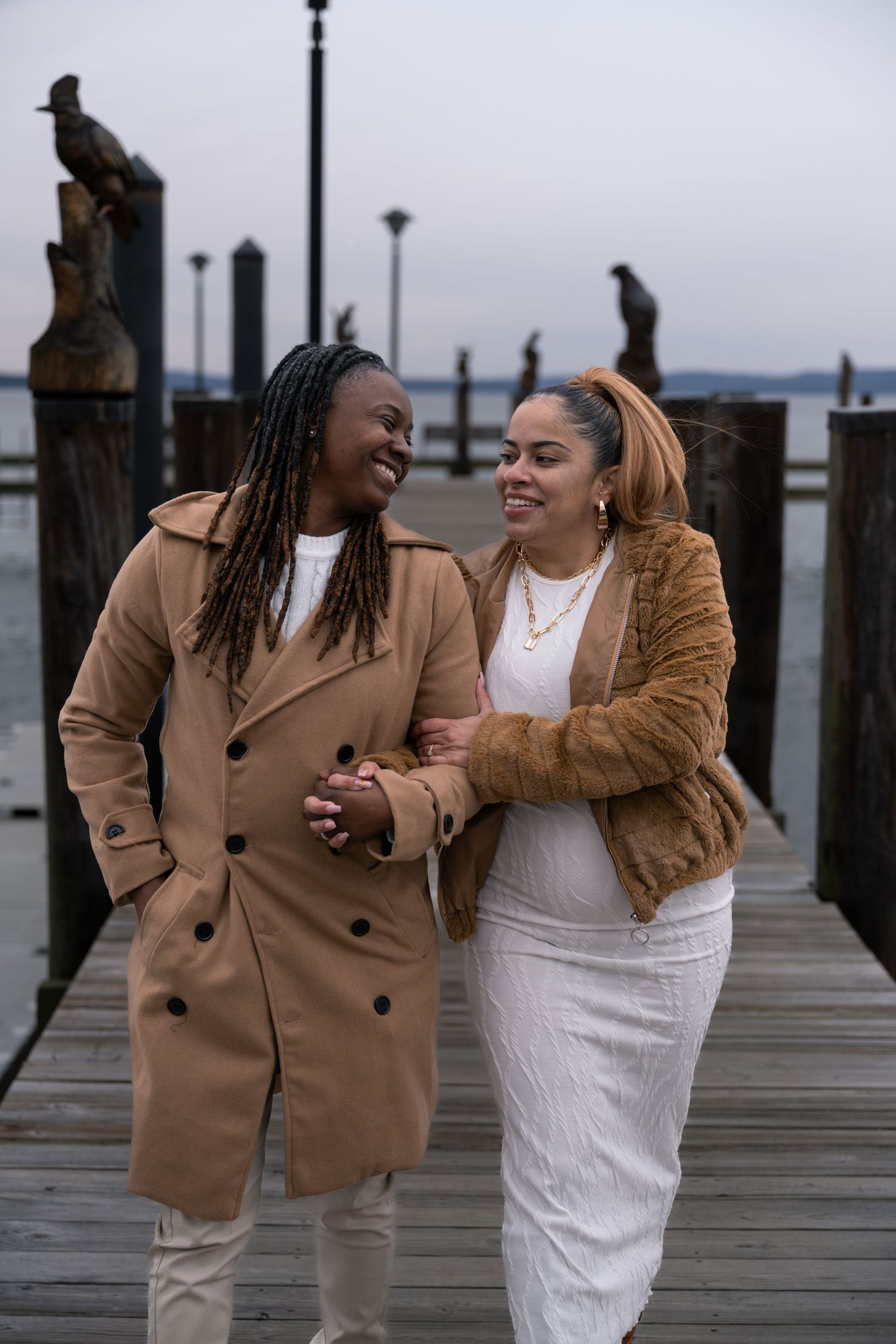 Couple laughing together on a wooden pier wearing brown coats and white outfits.