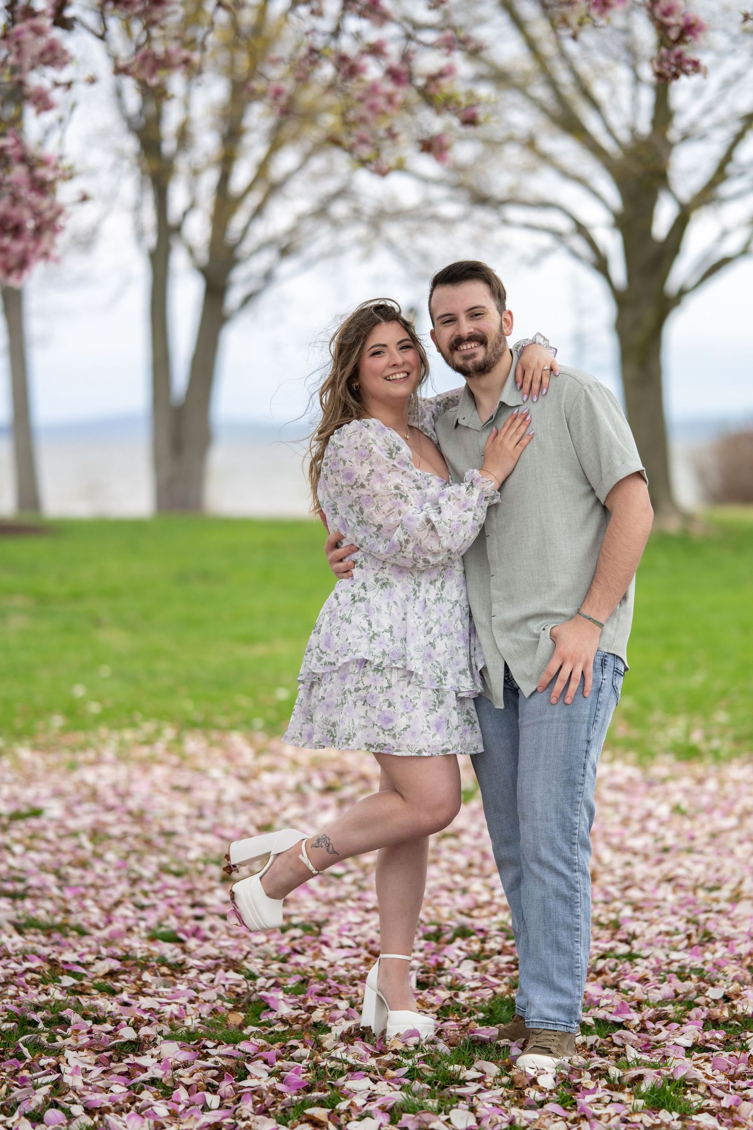 A couple shares a playful moment together among pink cherry blossom petals scattered on green grass.