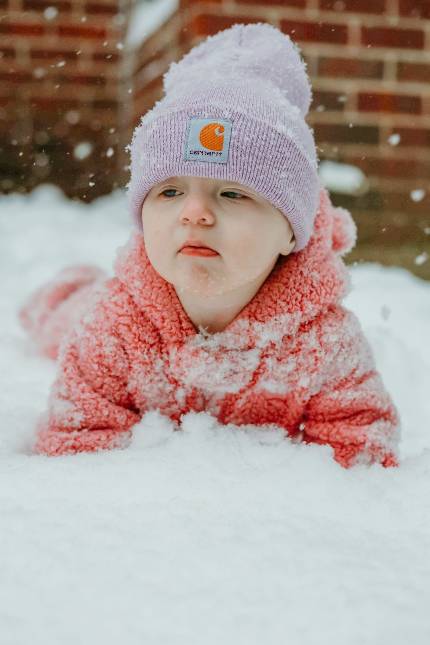 Small figure in a lavender Carhartt beanie and coral snowsuit lies in fresh snow while snowflakes fall around them.