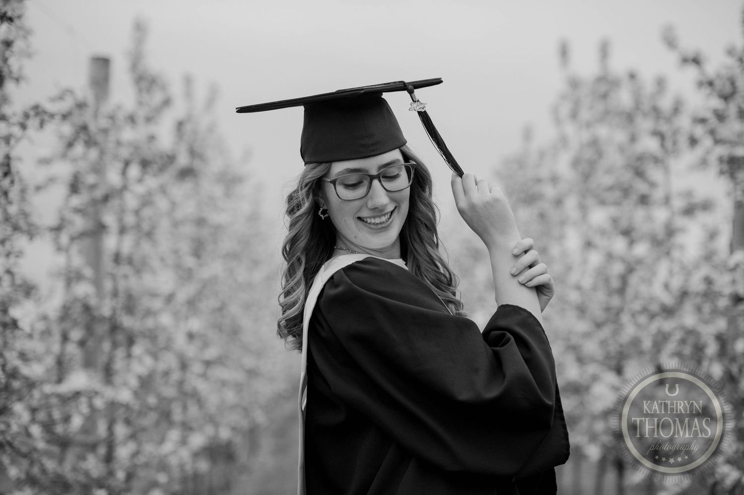 A graduate in cap, gown and glasses celebrates outdoors in black and white photography.
