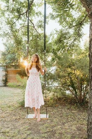 Woman in floral dress standing on wooden swing under trees with golden sunset light in background.