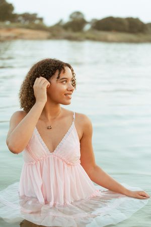 Young woman in pink lace dress smiling while sitting in calm water, with curly hair and a delicate necklace.