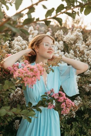 Woman in light blue dress and glasses posing among blooming pink and white flowers in golden hour light.