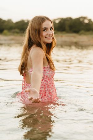 Young woman in pink floral dress smiling while standing in a lake during golden hour.
