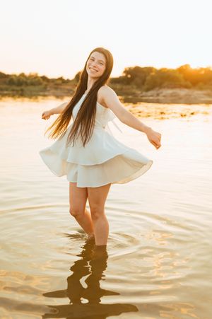 A young woman in a white dress twirls joyfully in shallow water during golden hour at the beach.