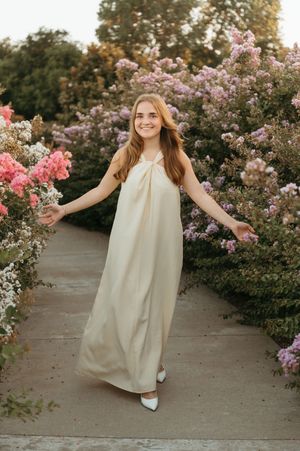 Young woman in cream maxi dress smiling among blooming pink and purple flower bushes on a garden path.