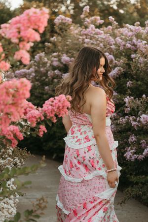 Woman in pink floral tiered dress posing among blooming pink and purple flower bushes in a garden.
