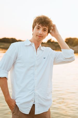 Young person in white button-up shirt poses against golden sunset by the water.