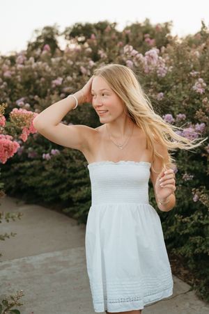 Young blonde woman in white strapless dress smiling outdoors among pink and purple flowering bushes.