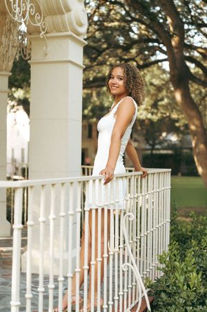 Young woman in white dress smiling on elegant white porch railing surrounded by lush green trees.
