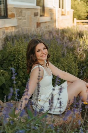 Woman in white summer dress and boots poses casually by rustic wooden fence in outdoor garden setting.