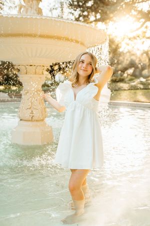 A person in a white dress sits on coastal rocks during golden hour sunset, with water and trees in the background.