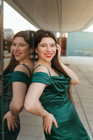 Woman in emerald green off-shoulder satin gown smiling beside mirror reflection, wearing gold earrings and red lipstick.