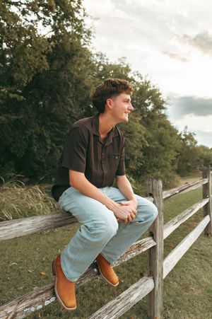 Person in light blue shirt and khaki pants relaxing on wooden dock by serene lake on cloudy day.