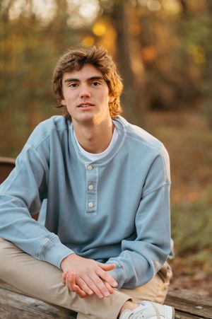 Person in light blue shirt and khaki pants relaxing on wooden dock by serene lake on cloudy day.