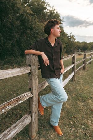 Young man in dark shirt and light jeans leaning on rustic wooden fence in rural setting at dusk.