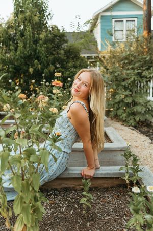 A dreamy figure in a light vintage dress sits in a wildflower meadow at golden hour with a wistful expression.