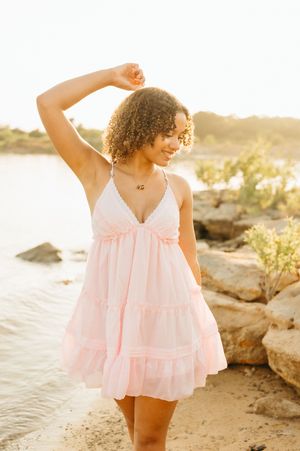 Young woman in pink tiered mini dress posing on rocky beach at golden hour sunset.