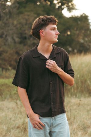 Young man in dark button-up shirt and light jeans standing in a field with trees in background.